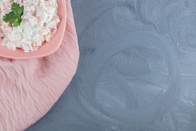 Pink bowl of olivier salad garnished with parsley leaves on pink tablecloth on marble table.