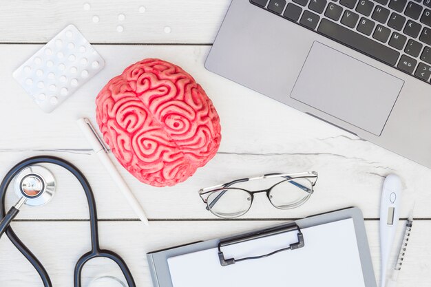 Pink brain model; pills; stethoscope; pen; eyeglasses; clipboard; thermometer; syringe and laptop on wooden desk