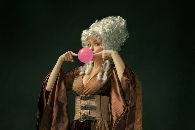 Free photo pink bubble gum. portrait of medieval young woman in brown vintage clothing on dark background. female model as a duchess, royal person. concept of comparison of eras, modern, fashion, beauty.