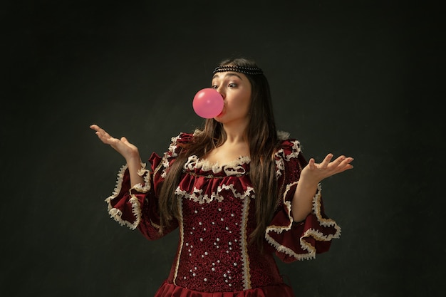 Pink bubble gum. Portrait of medieval young woman in red vintage clothing standing on dark background.