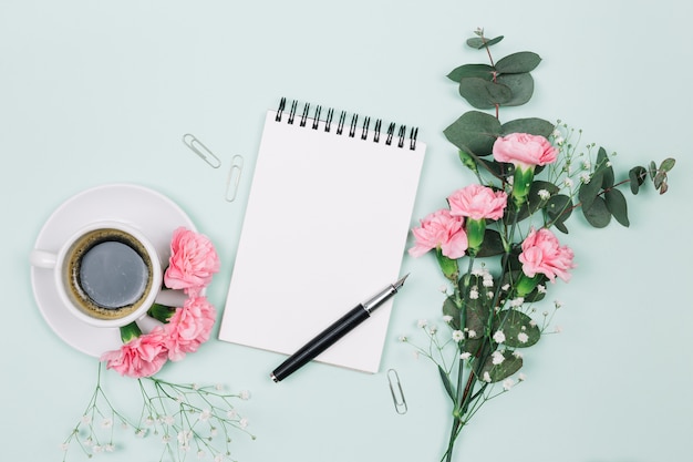 ピンクのカーネーションとジプソフィラの花とコーヒーカップ。スパイラルメモ帳と青い背景に万年筆