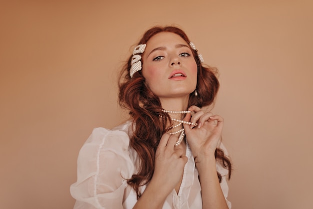 Pink-cheeked redhead woman plays with her pearl necklace. Lady in classic white blouse looking down at camera.