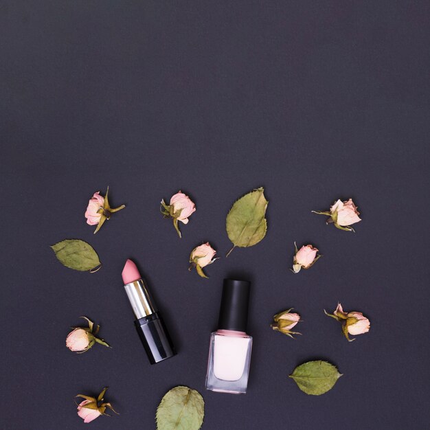 Pink lipstick and nail varnish bottle surrounded with pink rose buds and leaves on black background