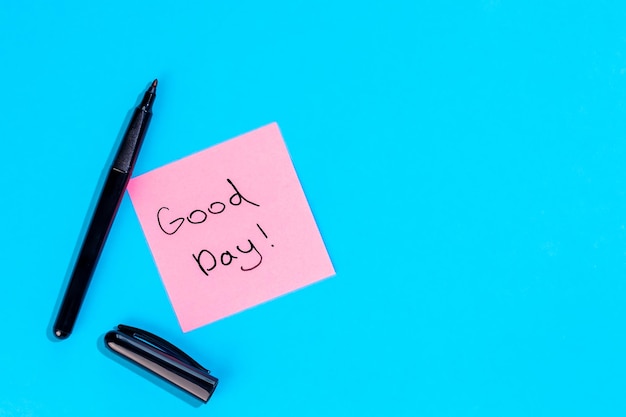 Free photo pink note with inscription good day and marker on blue background flat lay