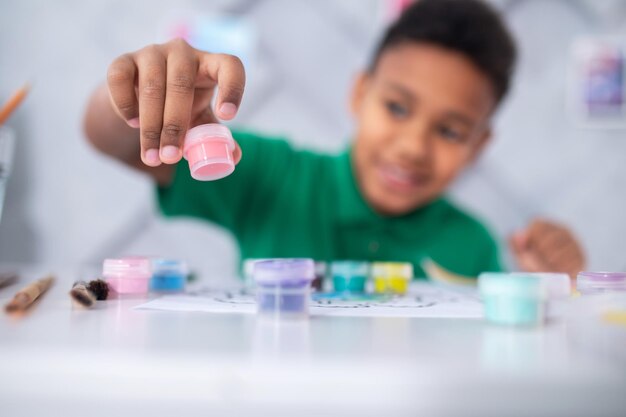 Pink paint. Cheerful dark-skinned boy in green tshirt stretching his hand with tube of pink paint for drawing at camera sitting at table in bright room