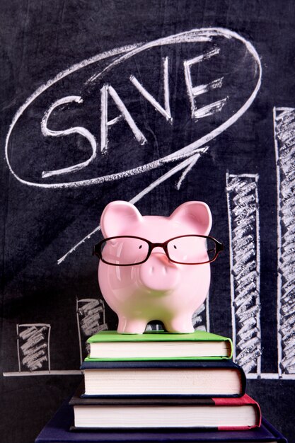 Розовая копилка с стеклами стоя на книгах рядом с классн классным с диаграммой роста сбережений.