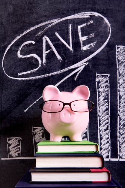 Pink piggy bank with glasses standing on books next to a blackboard with savings growth chart.  