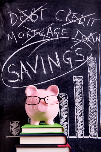 Free photo pink piggy bank with glasses standing on books next to a blackboard with savings message.