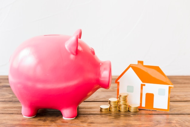 Pink piggybank near stacked coins and house on wooden desk