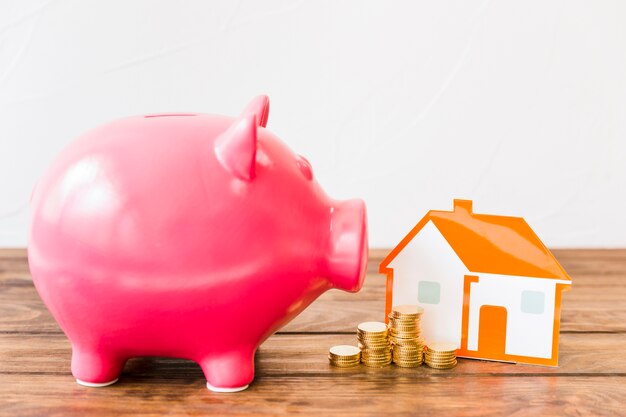 Pink piggybank near stacked coins and house on wooden desk