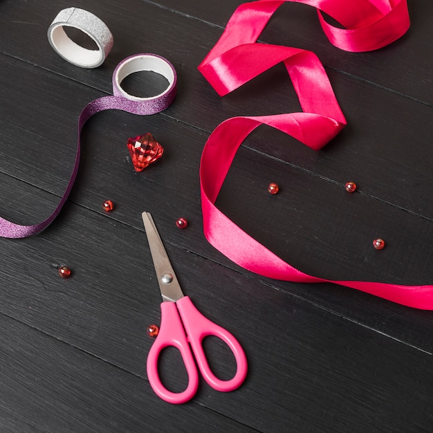 Free photo pink ribbon; colorful sticky tape; pearls; diamond and scissor on black wooden table