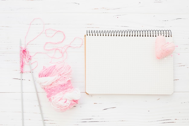 Pink yarn with crochet beside notepad and heart on wooden background