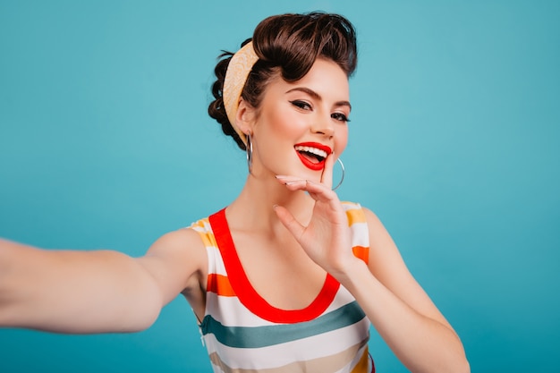 Free photo pinup girl with bright makeup posing on blue background. studio shot of brunette woman taking selfie.