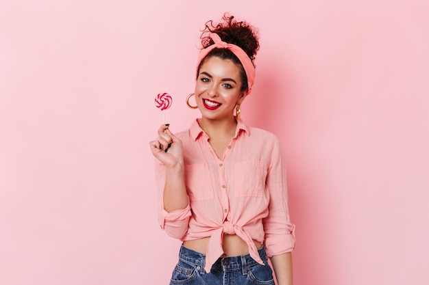 Pinup woman in stylish headband and pink shirt posing on isolated space and holding lollipop.