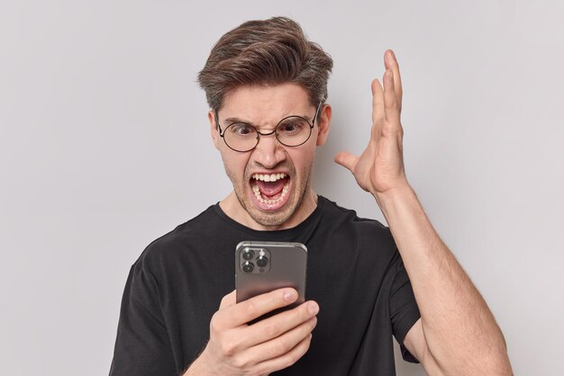 Pissed off annoyed man screams angrily keeps palm raised stares at smartphone being outraged after rough conversation wears round spectacles casual black t shirt isolated over white background