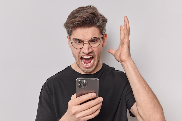 Разъяренный раздраженный мужчина сердито кричит, держит ладонь поднятой и смотрит на смартфон, возмущенный после грубого разговора, носит круглые очки, повседневную черную футболку, изолированную на белом фоне.
