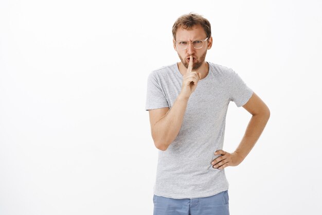 Pissed serious-looking strict and annoyed attractive guy with bristle in glasses frowning making shush gesture with index finger over mouth warning not spread secret