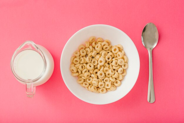 Pitcher of milk and bowl of cereals and spoon on the red background