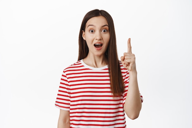 Pitching an idea. Excited young woman has suggestion, pointing up, raising finger and say suggestion, looking amazed, telling big news, standing over white background