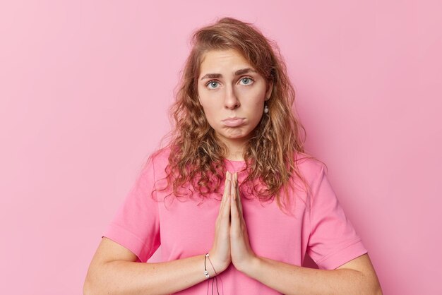 Pity sad blue eyed woman quivers lips with upset expression presses palms together has hopeless begging expression asks for apologize or favor wears casual t shirt isolated over pink background.