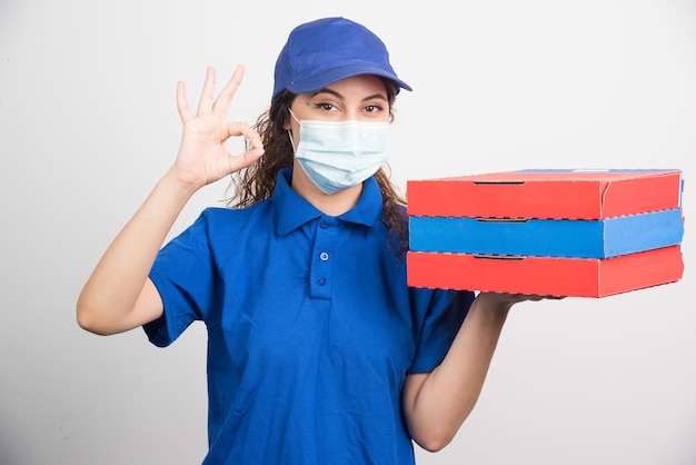 Pizza delivery girl holding three boxes with medical facemask showing ok gesture on white