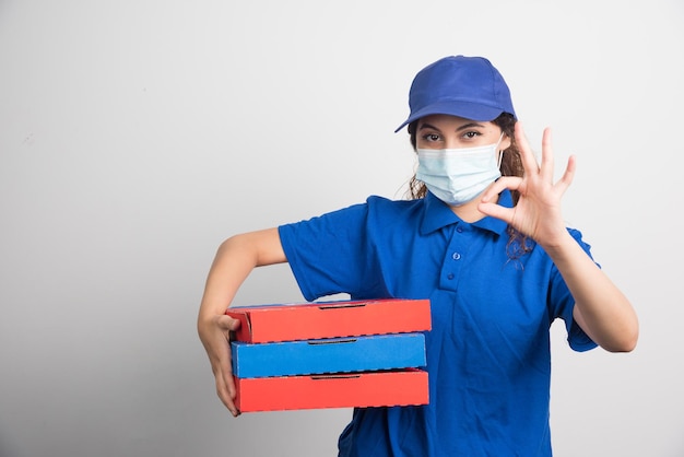 Pizza delivery girl holding three boxes with medical facemask and shows ok gesture on white