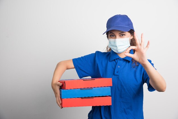 Доставка пиццы держит три коробки с медицинской маской и показывает жест на белом