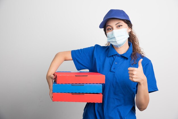 Pizza delivery girl holding three boxes with medical facemask and shows thumb up on white