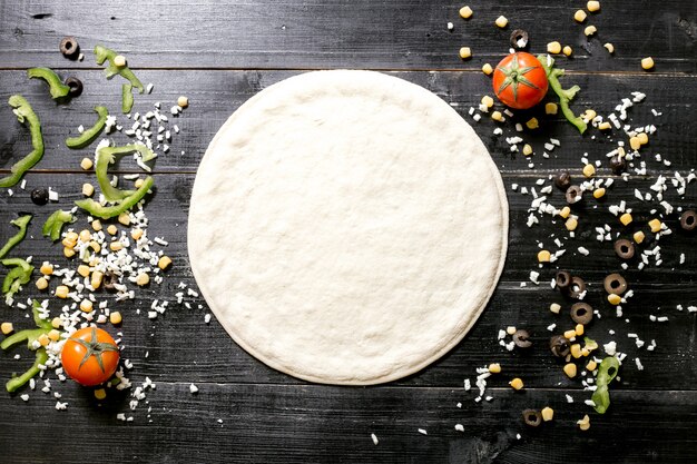 Pizza dough next to cheese sprinkles olive corn tomato and bell pepper on black wooden background