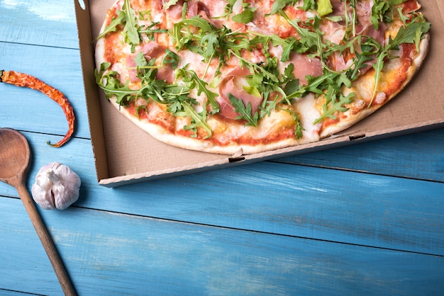 Пицца с листьями бекона и рукколы в картонной коробке с красным перцем чили и луковицей чеснока над деревянным столом