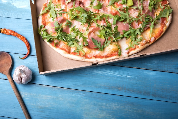 Pizza with bacon and arugula leaves in cardboard box with red chili and garlic bulb over wooden table