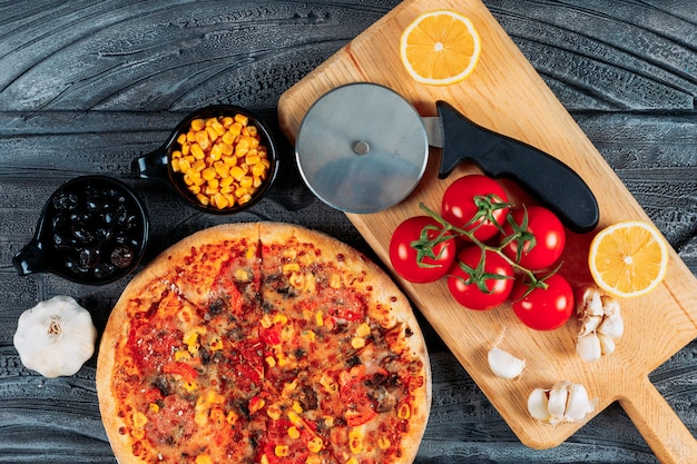 Pizza with garlic, tomatoes, a lemon, olives, corn and a pizza cutter top view on a dark wooden background