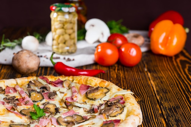 木製​の​テーブル​、​トマト​、​マッシュルーム​、​その他​の​野菜​を​背景​に​ハム​、​タマネギ​、​ピクルス​の​ピザ