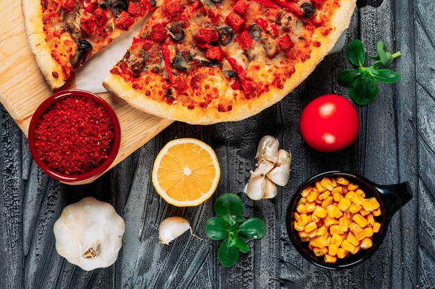 Pizza with a tomato, slices of garlic and lemon, chili pepper, corn and mint leaves in a pizza board on gray wooden background, high angle view.