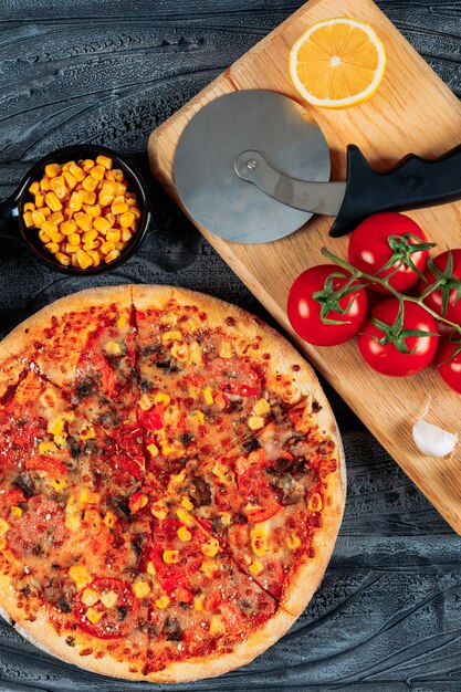 Pizza with tomatoes, a slice of lemon and garlic, corn and a pizza cutter high angle view on a dark wooden background