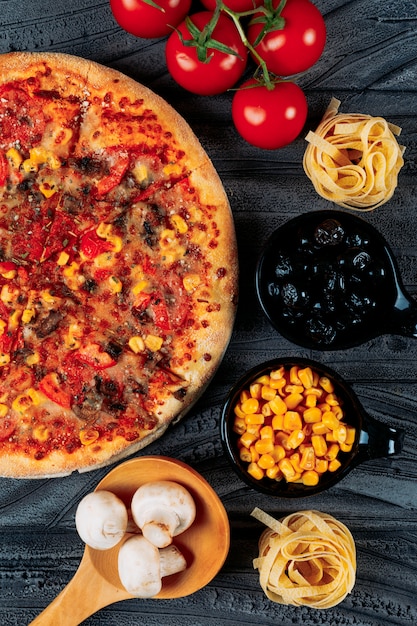 Pizza with tomatoes, spaghetti, corn, olives, mushrooms close-up on a dark background