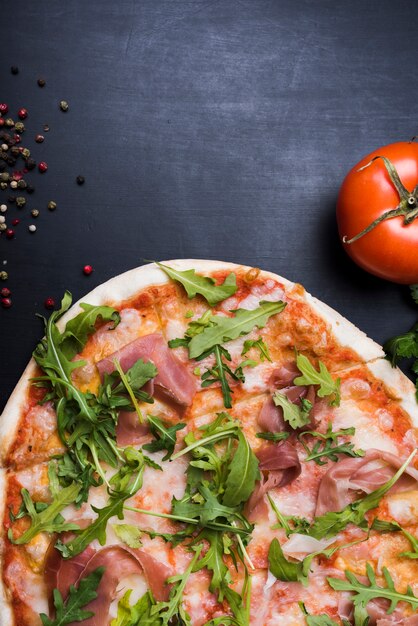 Pizza with topping of bacon and arugula leaves near juicy tomato and black pepper on textured black surface