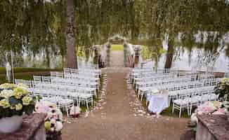 Foto gratuita luogo per la cerimonia di nozze con arco nuziale decorato con fiori e sedie bianche su ciascun lato dell'arco all'aperto. preparazione per la cerimonia di matrimonio all'aperto vicino al lago.