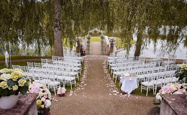 Place for wedding ceremony with wedding arch decorated with flowers and white chairs on each side of archway outdoors. Preparation for wedding ceremony outdoors near lake.