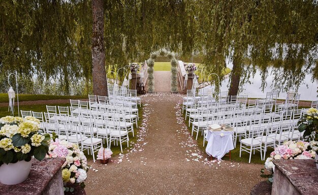 Place for wedding ceremony with wedding arch decorated with flowers and white chairs on each side of archway outdoors. Preparation for wedding ceremony outdoors near lake.