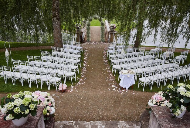 Place for wedding ceremony with wedding arch decorated with flowers and white chairs on each side of archway outdoors. Preparation for wedding ceremony outdoors near lake.