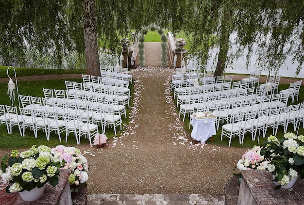 Место для свадебной церемонии со свадебной аркой, украшенной цветами и белыми стульями по обе стороны арки на открытом воздухе. Подготовка к свадебной церемонии на открытом воздухе у озера.