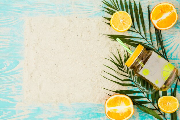 Plant leaves near glass of drink and oranges with sand on board