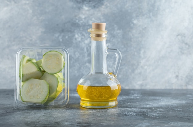 Plastic container full with zucchini pieces and bottle of oil over grey background.