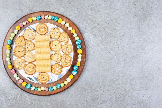 Тарелка ассорти печенья в окружении конфет попкорна на деревянной доске на мраморном фоне. Фото высокого качества