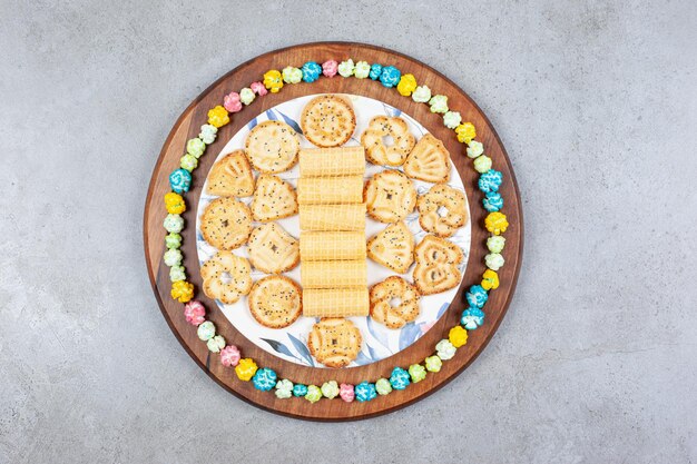 Plate of assorted cookies encircled by popcorn candy on wooden board on marble surface.