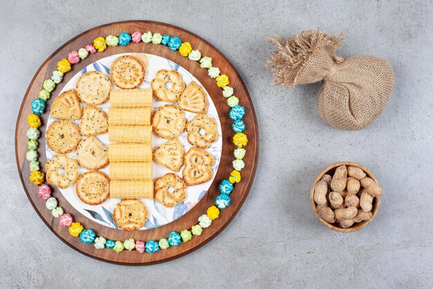 Plate of assorted cookies encircled by popcorn candy on wooden board next to a sack and a bowl of peanuts on marble surface.