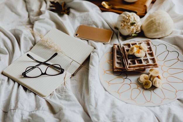 Plate of breakfast waffle with banana topping on a white bed next to a diary and a phone