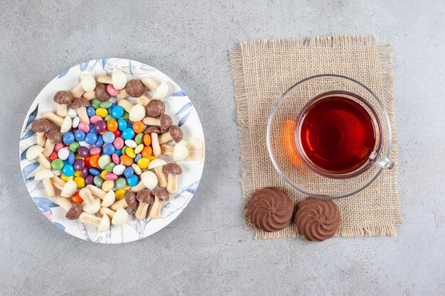 Тарелка конфет и шоколадных грибов рядом с чашкой чая и двумя печеньями на мраморной поверхности