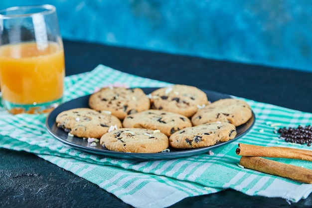 Free photo a plate of chocolate chips cookies and a glass of orange juice on dark table with cinnamon